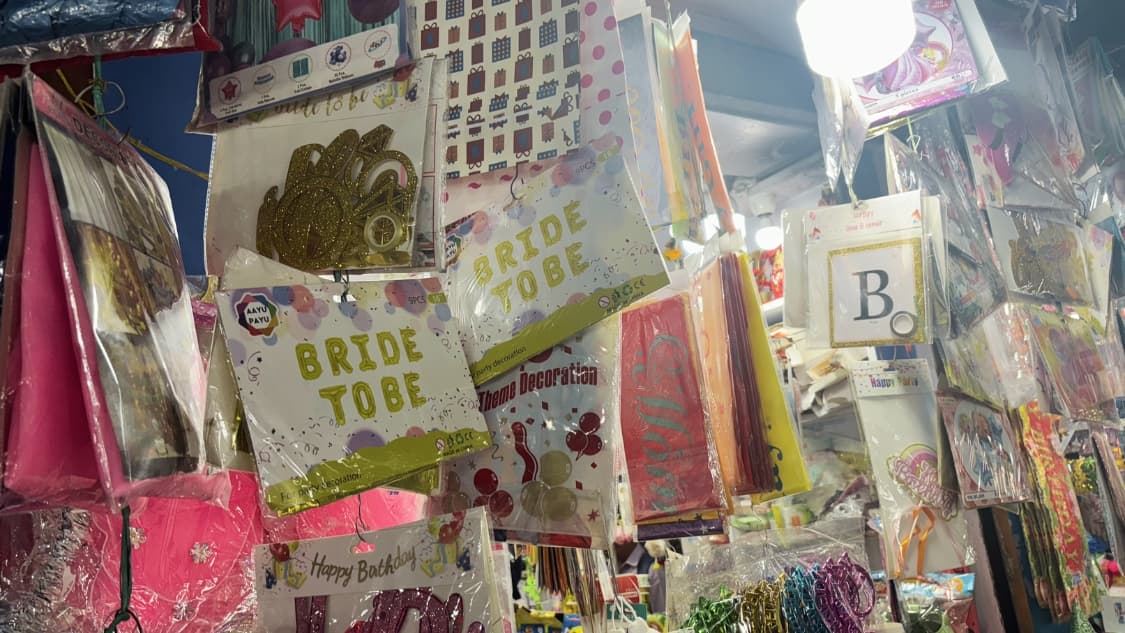 <p>A close-up of a party supply stall featuring "Bride to Be" banners, gold glitter cake toppers and various colorful gift-wrapping papers and decorative streamers.</p>