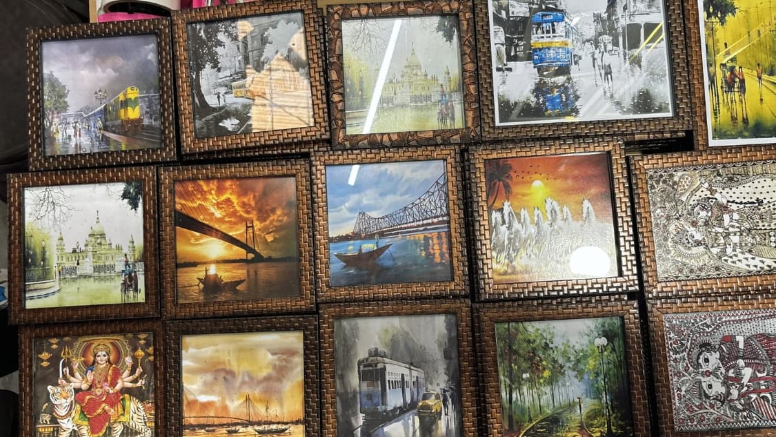 <p>A collection of framed miniature artworks showcasing iconic Kolkata landmarks, including the Howrah Bridge, yellow taxis, trams and traditional Madhubani-style paintings.</p>