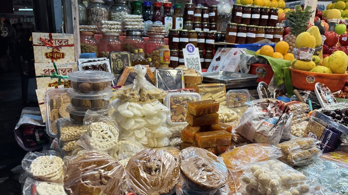 <p>A variety of traditional Bengali sweets and preserves, including white <em>batasa</em> (sugar drops), <em>aamshotto</em> (dried mango pulp), patali gur and jars of pickles and honey, neatly arranged at a market vendor's stall.</p>